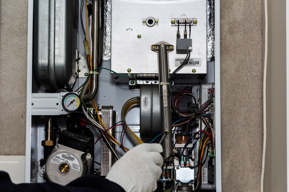 A gloved hand repairs or inspects the internal components of a gas boiler, with various wires, pipes, and mechanical parts visible inside the open unit&mdash;service like this is expertly handled by a skilled plumber in Boone County, MO.