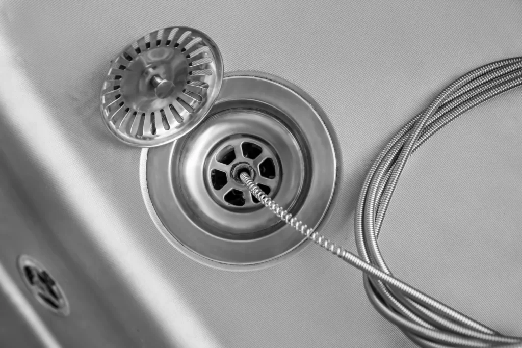A metal drain snake is inserted into a kitchen sink drain, with the strainer set aside—capturing a classic black and white scene of a plumber Boone County expert performing professional drain cleaning Boone County, MO.