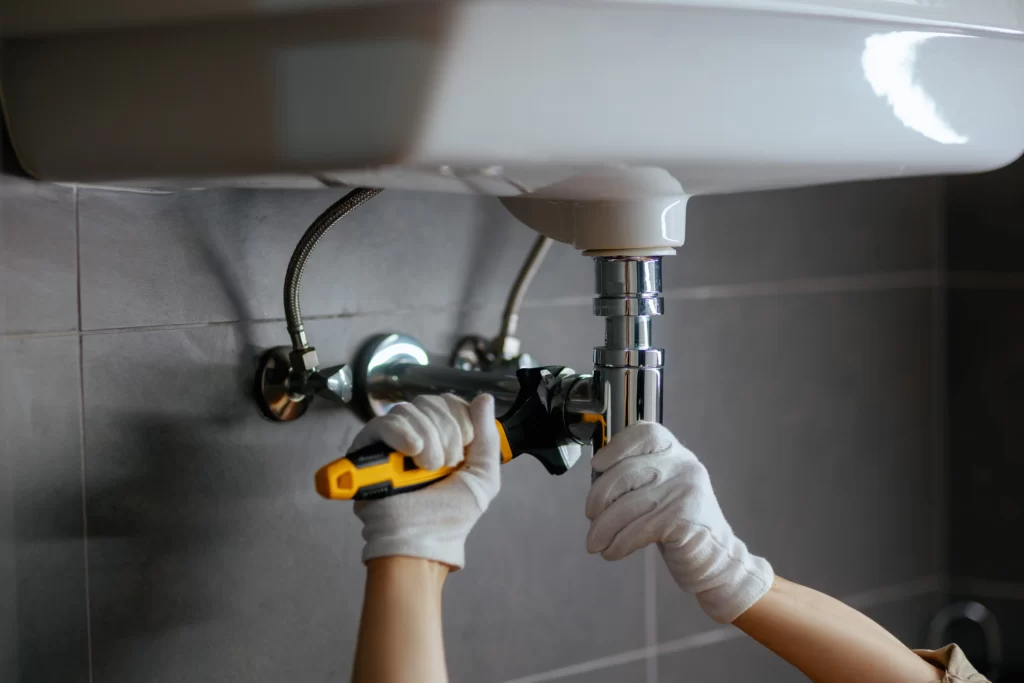 A plumber Boone County, MO, wearing white gloves uses tools to fix the plumbing under a sink, tightening a pipe beneath the sink in a bathroom or kitchen.