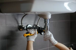 A plumber Boone County, MO, wearing white gloves uses tools to fix the plumbing under a sink, tightening a pipe beneath the sink in a bathroom or kitchen.