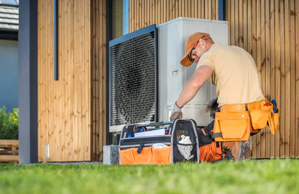 A technician wearing an orange cap and tool belt repairs an outdoor air conditioning unit beside a wooden building, with a tool bag open on the ground next to him—your trusted plumber Boone County for HVAC and drain cleaning needs.