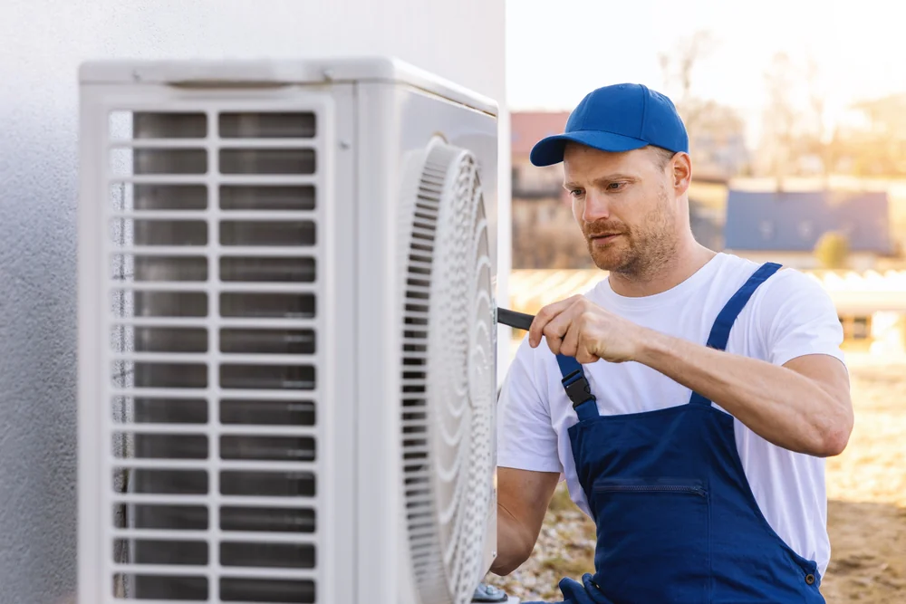 A technician in blue overalls and a cap uses a screwdriver to repair or install an outdoor air conditioning unit attached to a building wall, similar to the skilled drain cleaning Boone County professionals who tackle tough plumbing jobs.